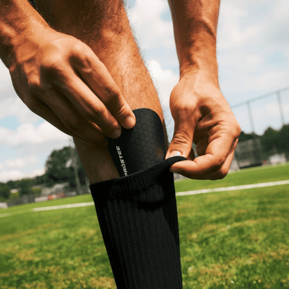 Speler doet Prostec scheenbeschermer, een voetbalaccessoire, met hexagonale patroon in zwarte sok op veld